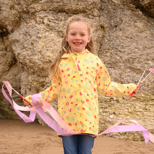 A child standing on a beach wearing a yellow waterproof jacket with a floral print, paired with blue jeans.