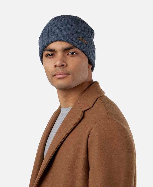 Man wearing a blue Barts Wilbert Turnup beanie in blue with a brown coat on a white background.