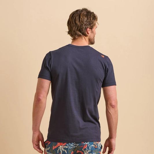 Back view of a man wearing a navy blue Brakeburn Island t-shirt with a small bird logo on the back against a beige background