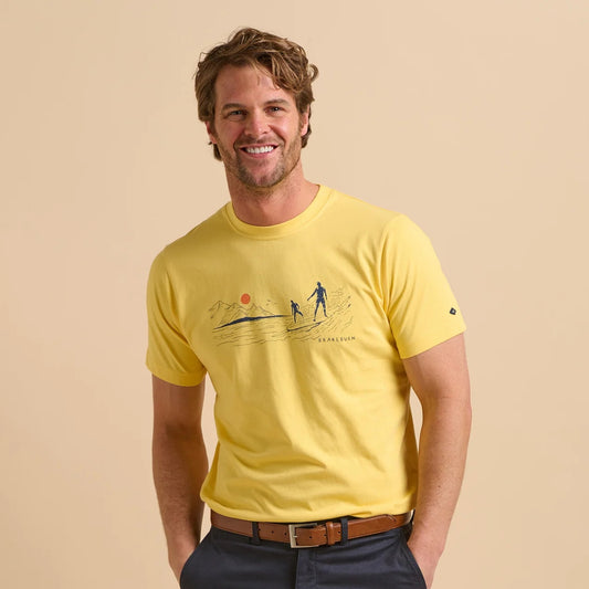 Front view of a man wearing a yellow Brakeburn t-shirt with a surf graphic design on a beige background