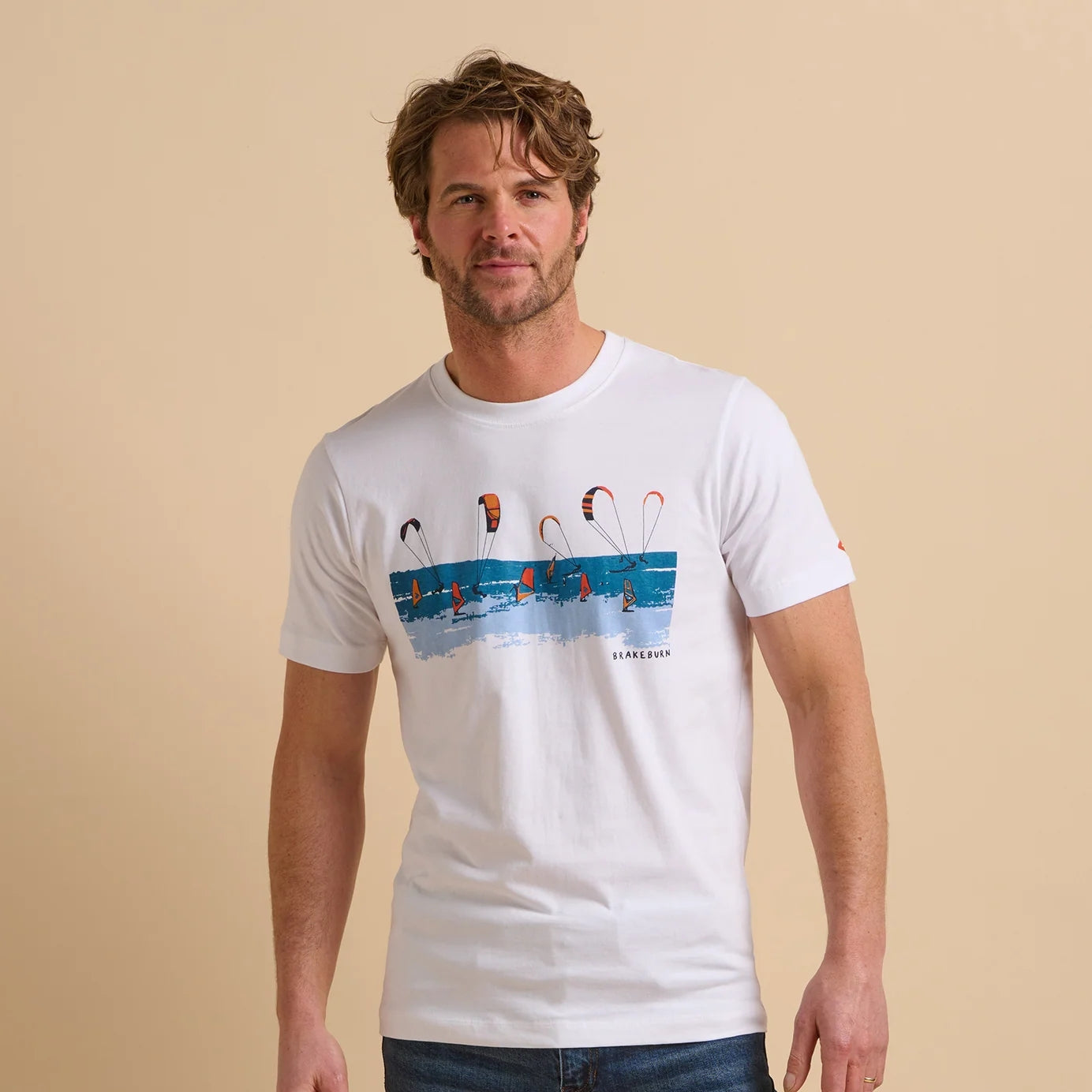 Front view of a man wearing a white short sleeve Brakebun tee with a sea, kite and windsurfer graphic design on a beige background