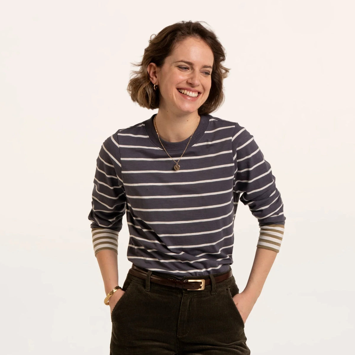 A woman wearing a Brakeburn long sleeve Exbury t-shirt in light navy with white stripes and contrasting mustard stripe cuffs ticked into dark jeans with a belt.