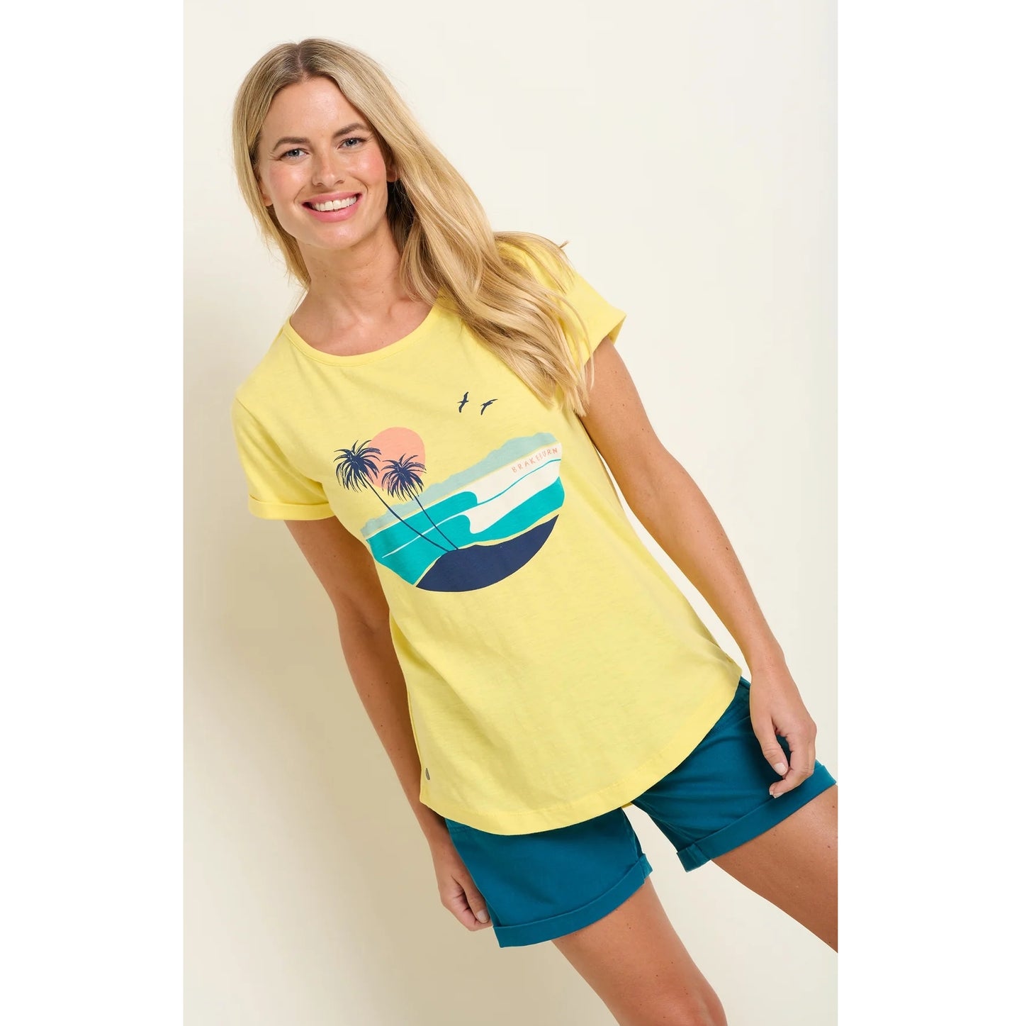 Front view of a Brakeburn womens yellow t-shirt with palm tree and ocean graphic worn with blue shorts