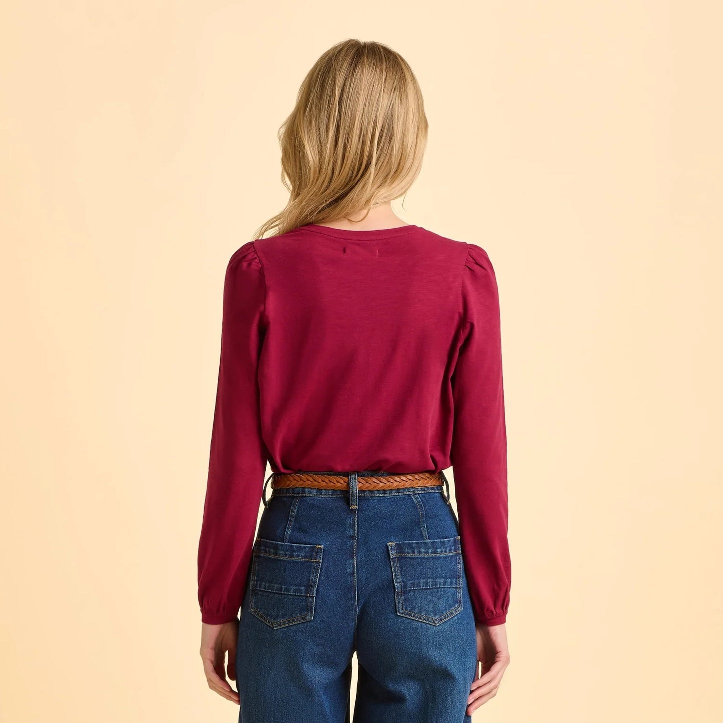 Back view of a woman wearing a burgundy red Brakeburn Janis long-sleeve t-shirt and blue jeans on a beige background