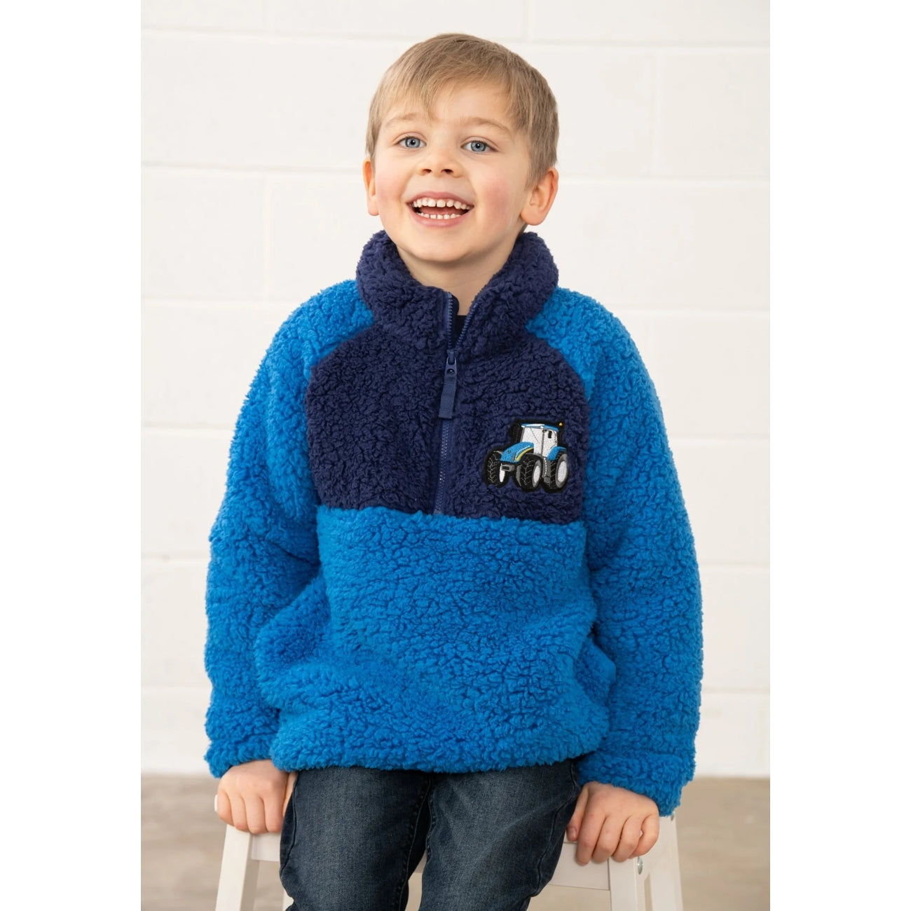 Child wearing a blue and navy Lighthouse Cadens fleece jumper with a tractor design on a white background