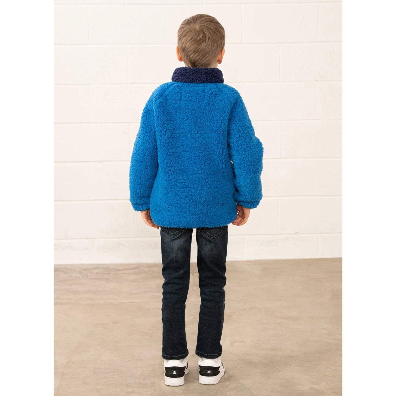 Back view of a child wearing a blue fluffy Lighthouse Cadens fleece standing against a white wall.