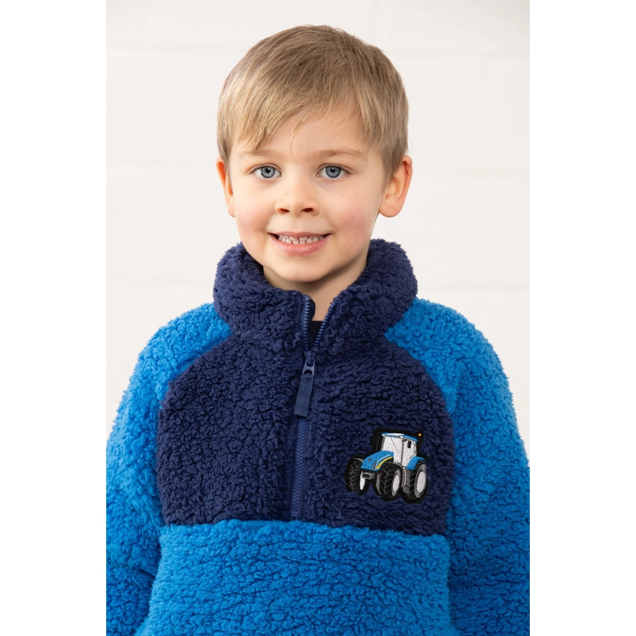 Child wearing a blue and navy Lighthouse Cadens fleece jumper with a tractor design on a white background