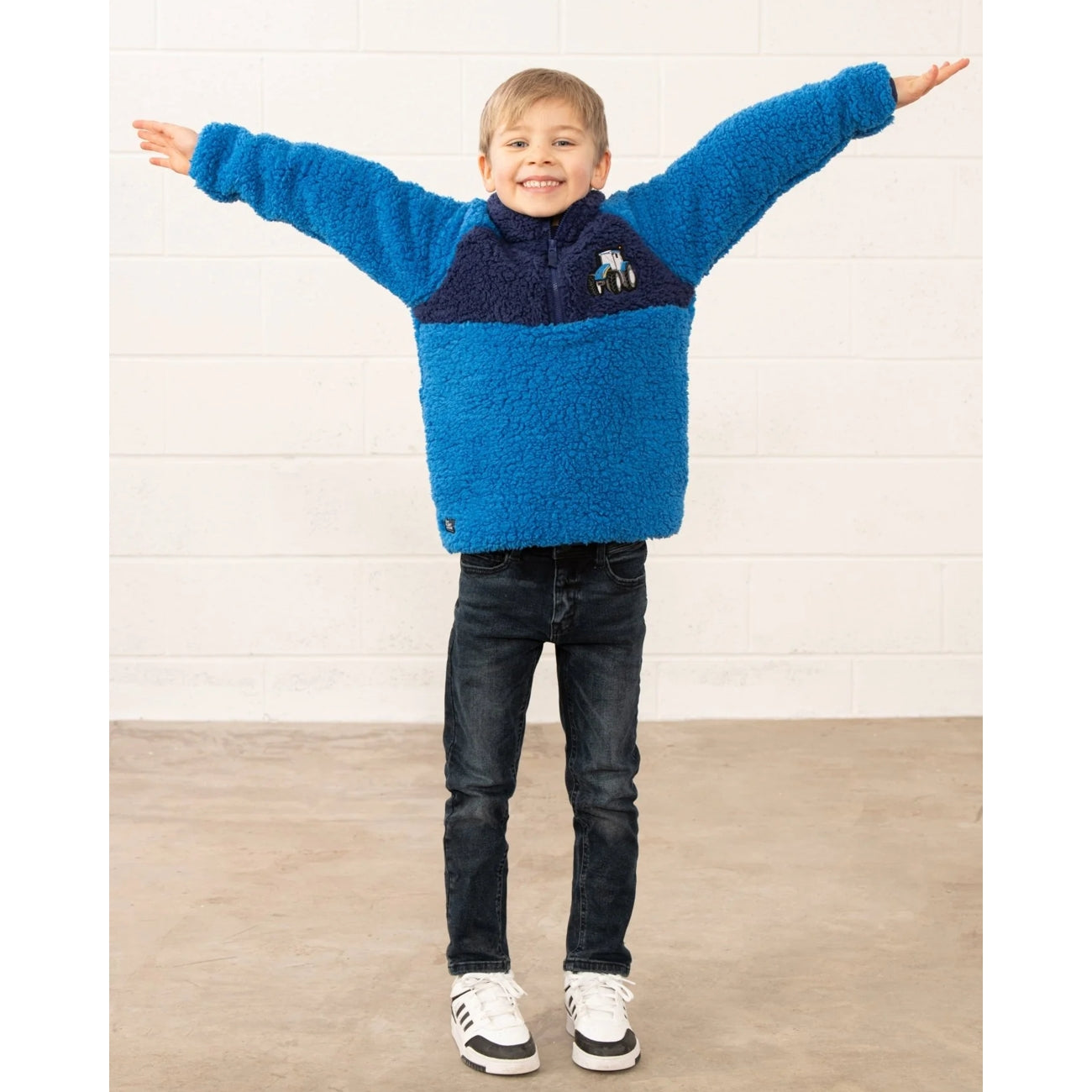Child wearing a blue Lighthouse Cadens fleece jumper with arms outstretched against a white wall.