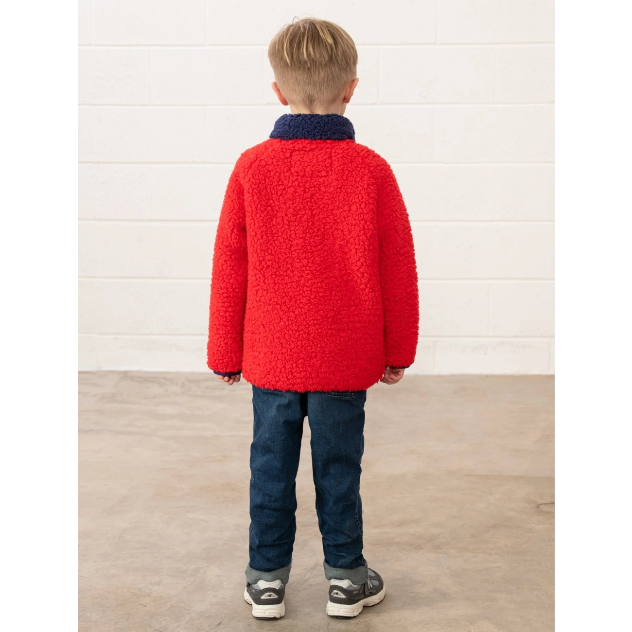 Back view of a child wearing a Lighthouse Caden red jumper and blue pants standing against a white wall.