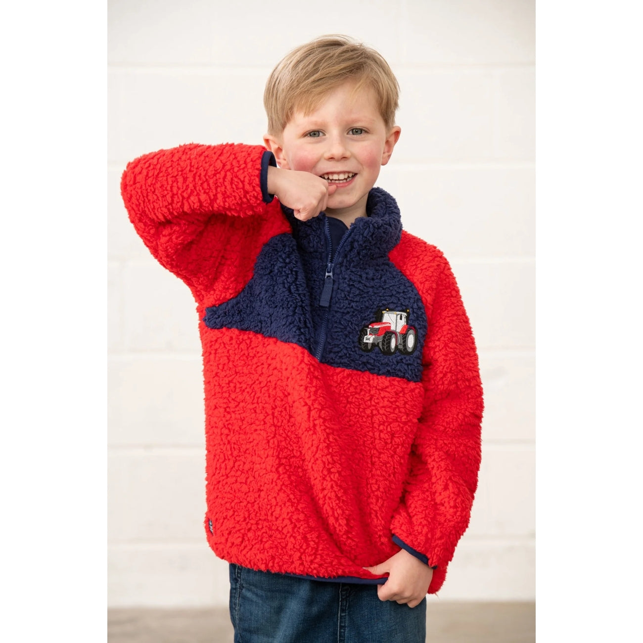 Front view of a child wearing a Lighthouse Caden red and navy fleece jumper with a tractor design on a white background