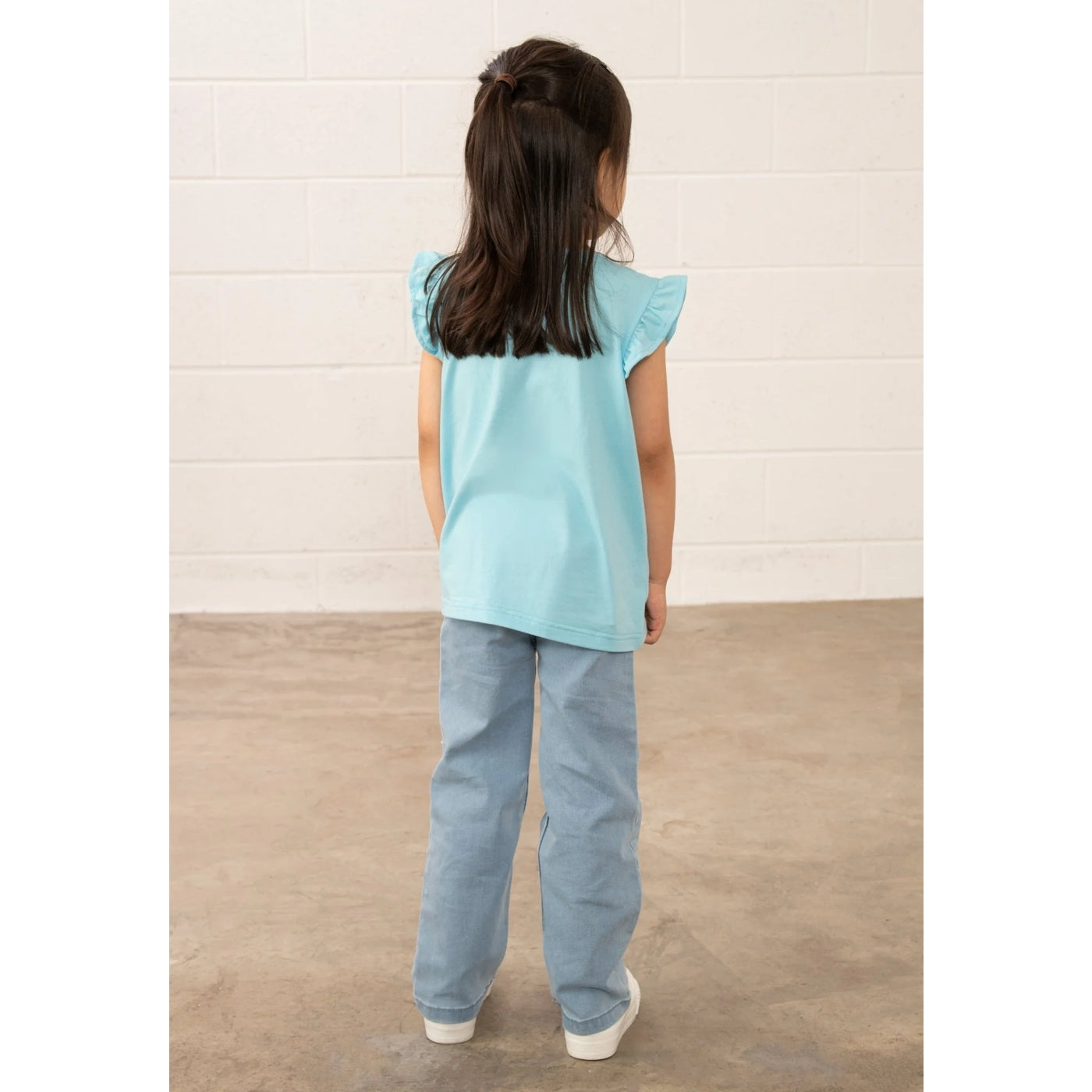 Child wearing a light blue Lighthouse top and jeans standing against a white wall.