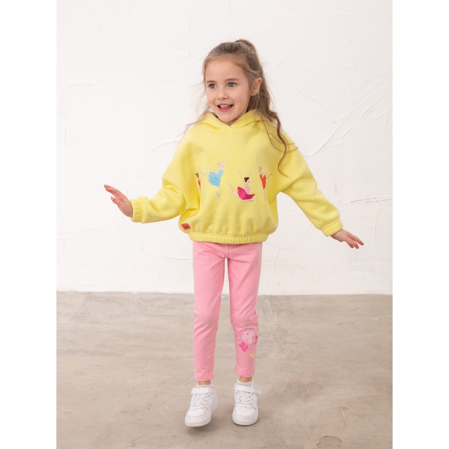Girl wearing a pair of Lighthouse kids Mollie leggings in Fluro Pink with a bow, heart and flower print on one leg with yellow hoodie.