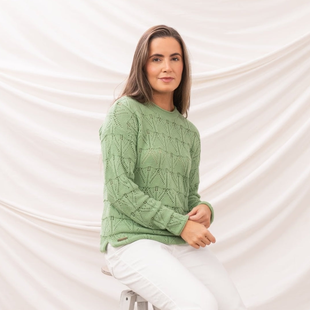 Side view of a woman wearing a Lighthouse Eden knitted jumper in Soft Sage Green with a geometric pointelle knit pattern with white trousers.
