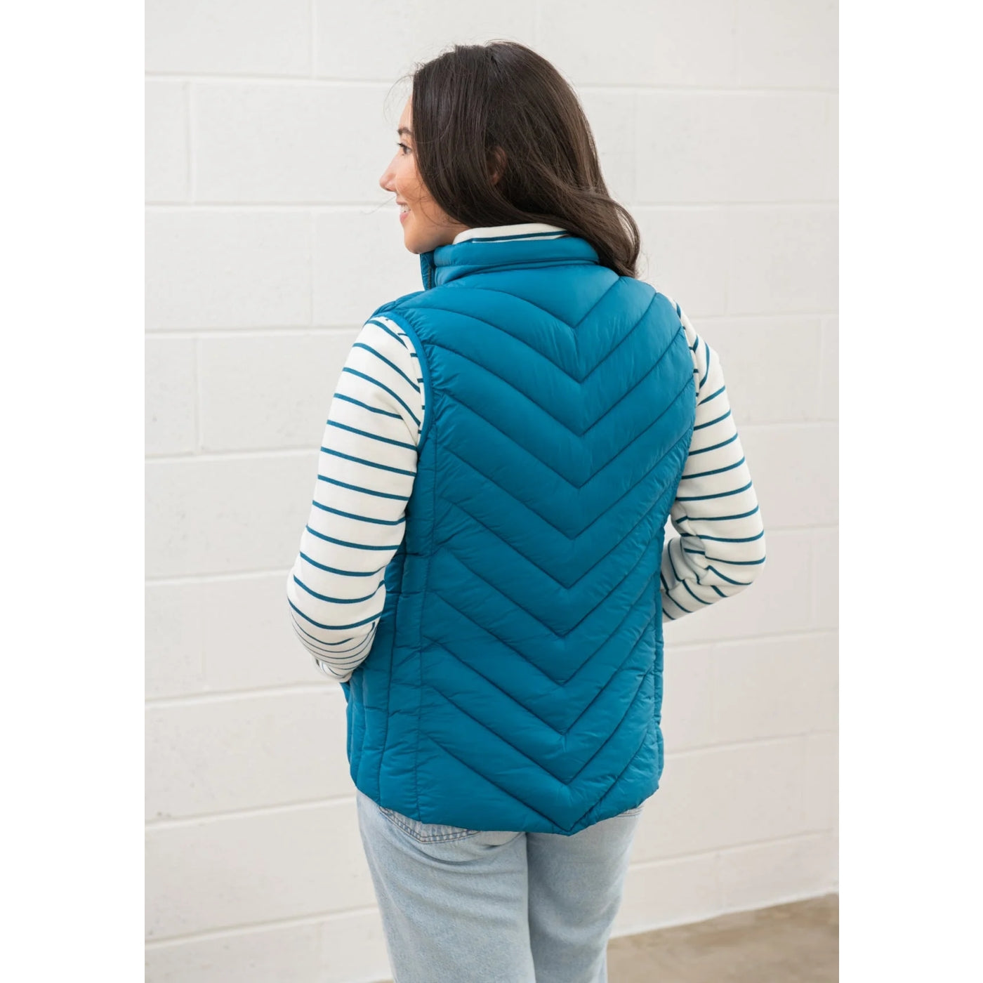 Back view of a woman wearing a Lighthouse Laurel teal blue quilted gilet against a white wall