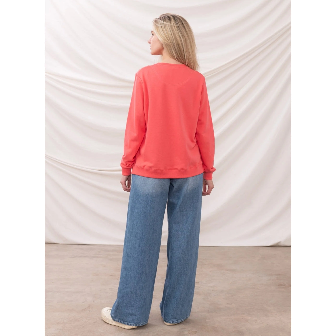 Woman wearing a coral Lighthouse sweatshirt and blue jeans standing against a plain white background