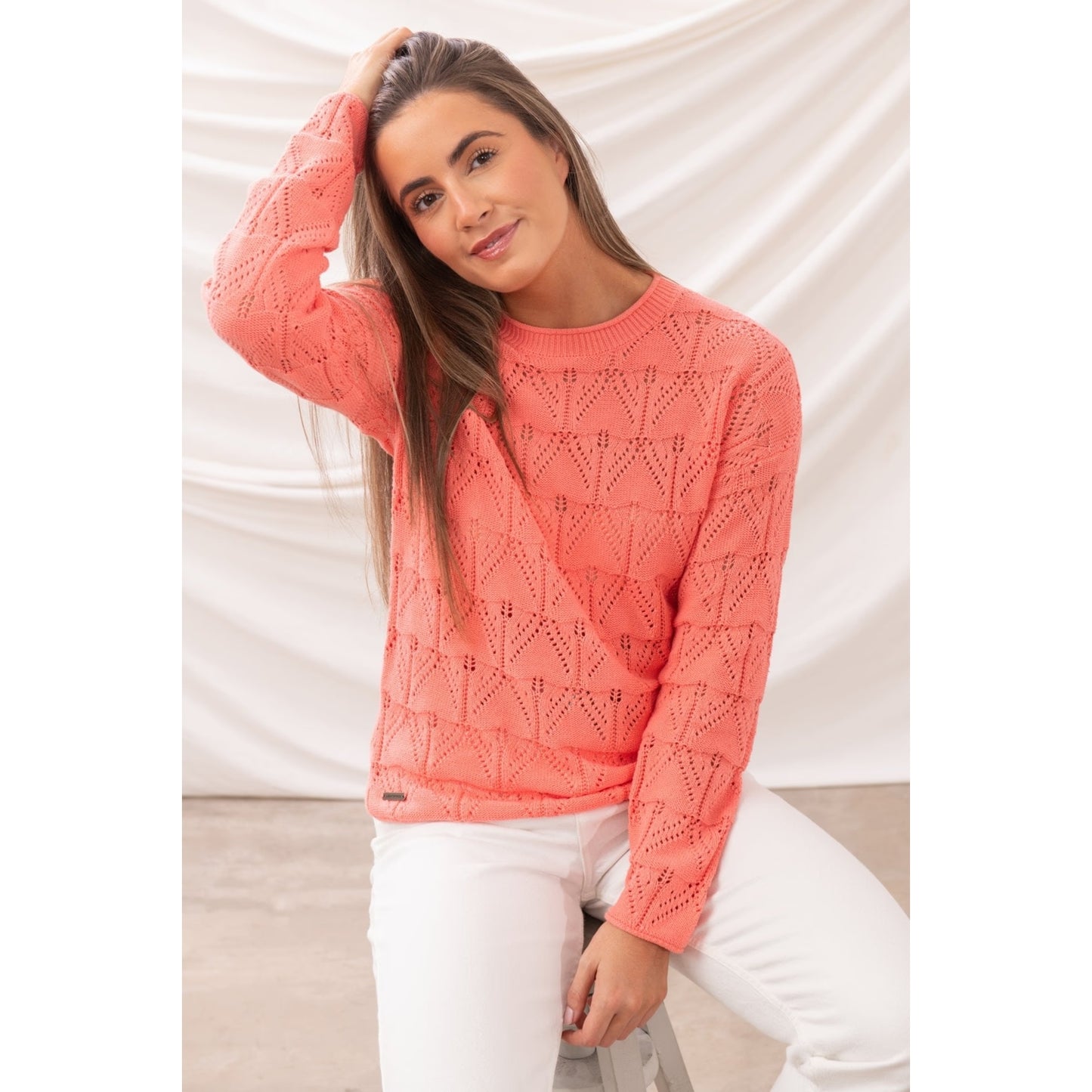 Woman wearing a Lighthouse knitted Eden jumper in Suga Coral with white trousers