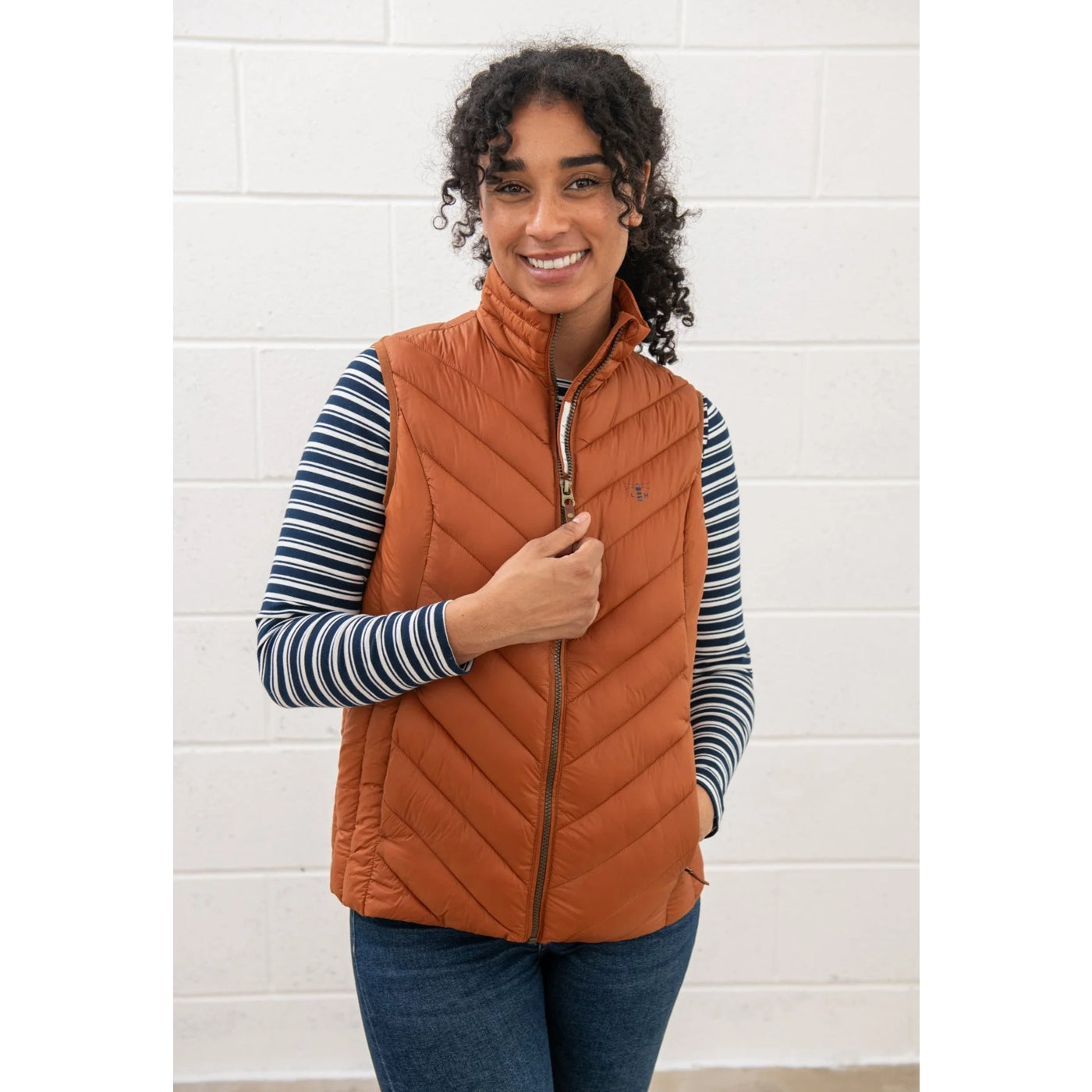 Woman wearing a Rust Orange Lighthouse Laurel padded gilet over a striped shirt against a white tiled wall.