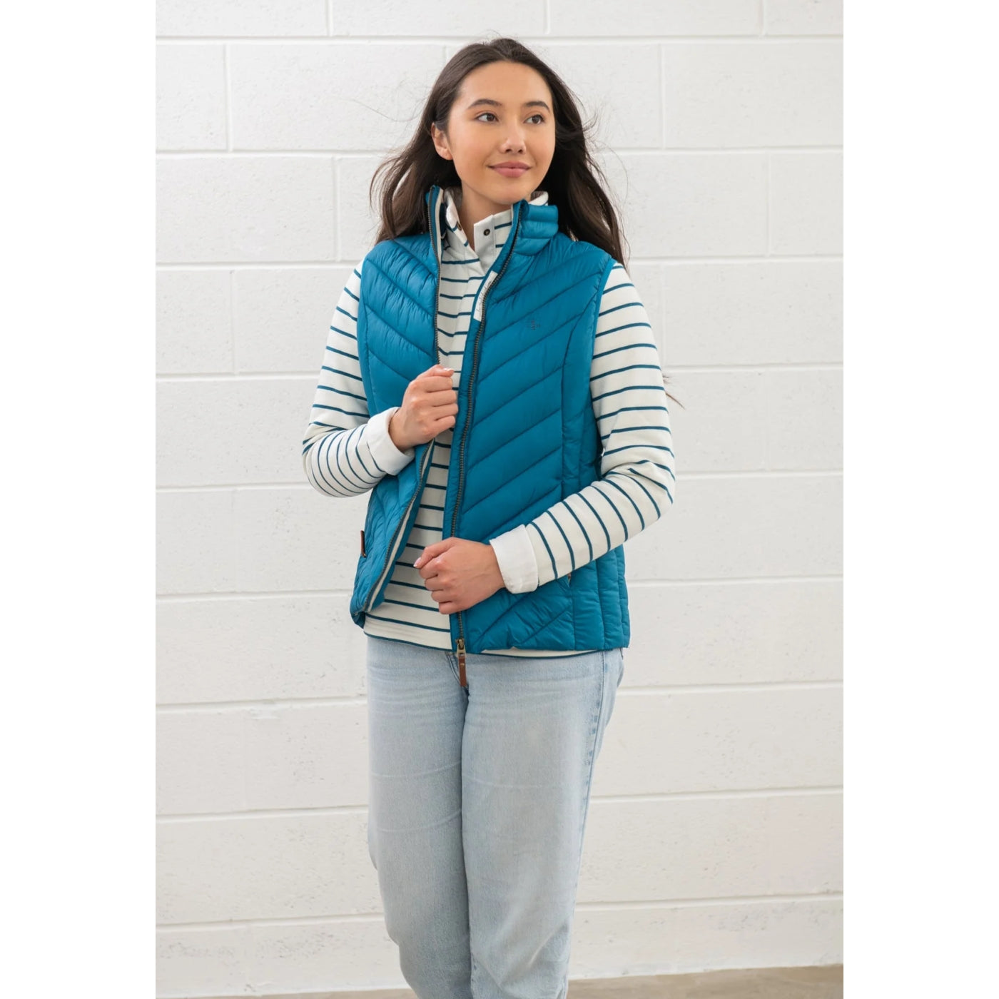 Woman wearing a Lighthouse Laurel puffer gilet in teal blue over a striped shirt against a white brick wall.