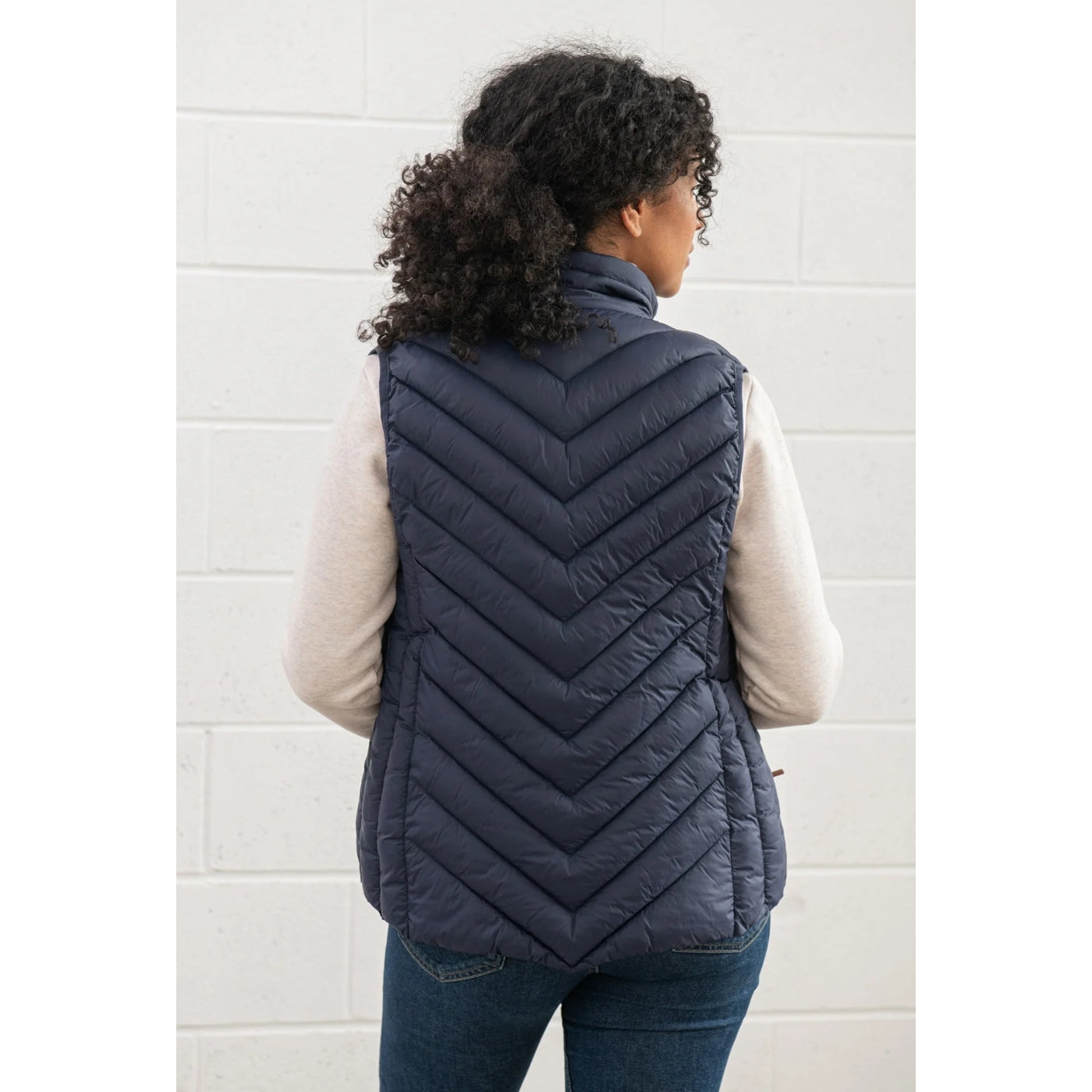 Back view of a woman wearing a navy quilted Lighthouse Laurel padded gilet against a white tiled wall