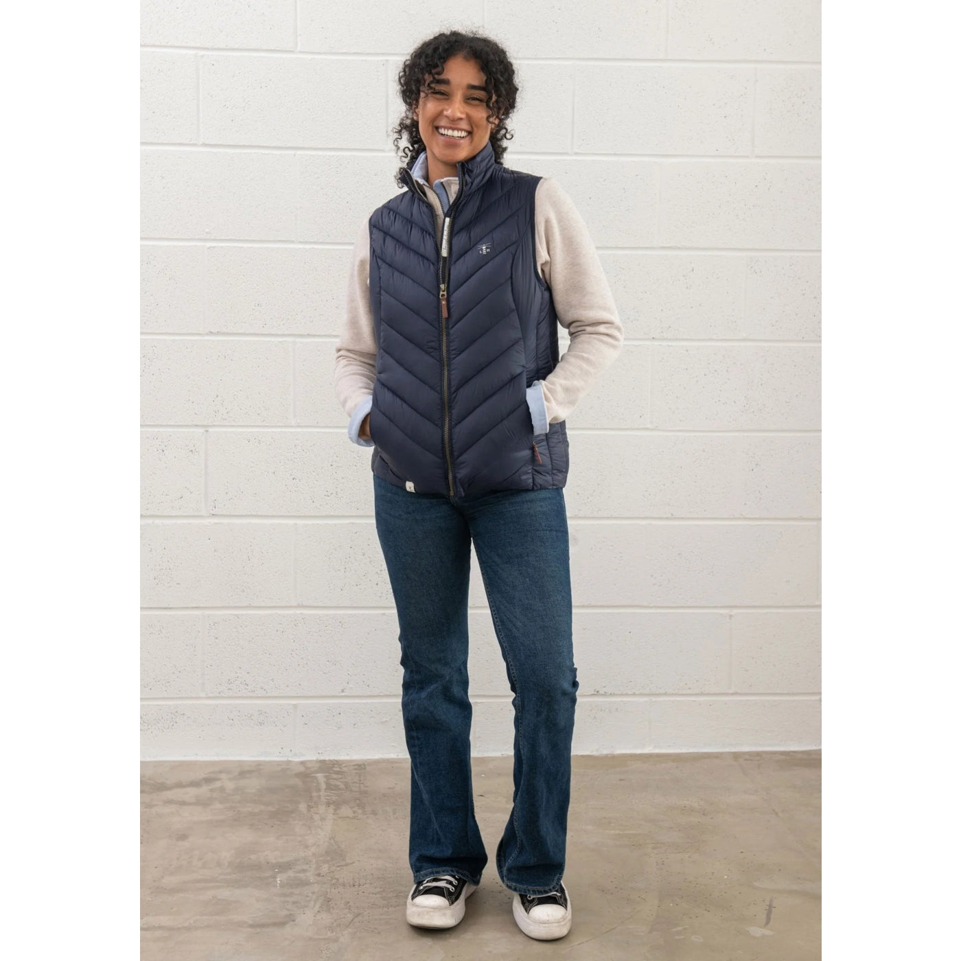 Woman wearing a navy padded Lighthouse Laurel gilet over a beige sweater and blue jeans against a light grey brick wall.