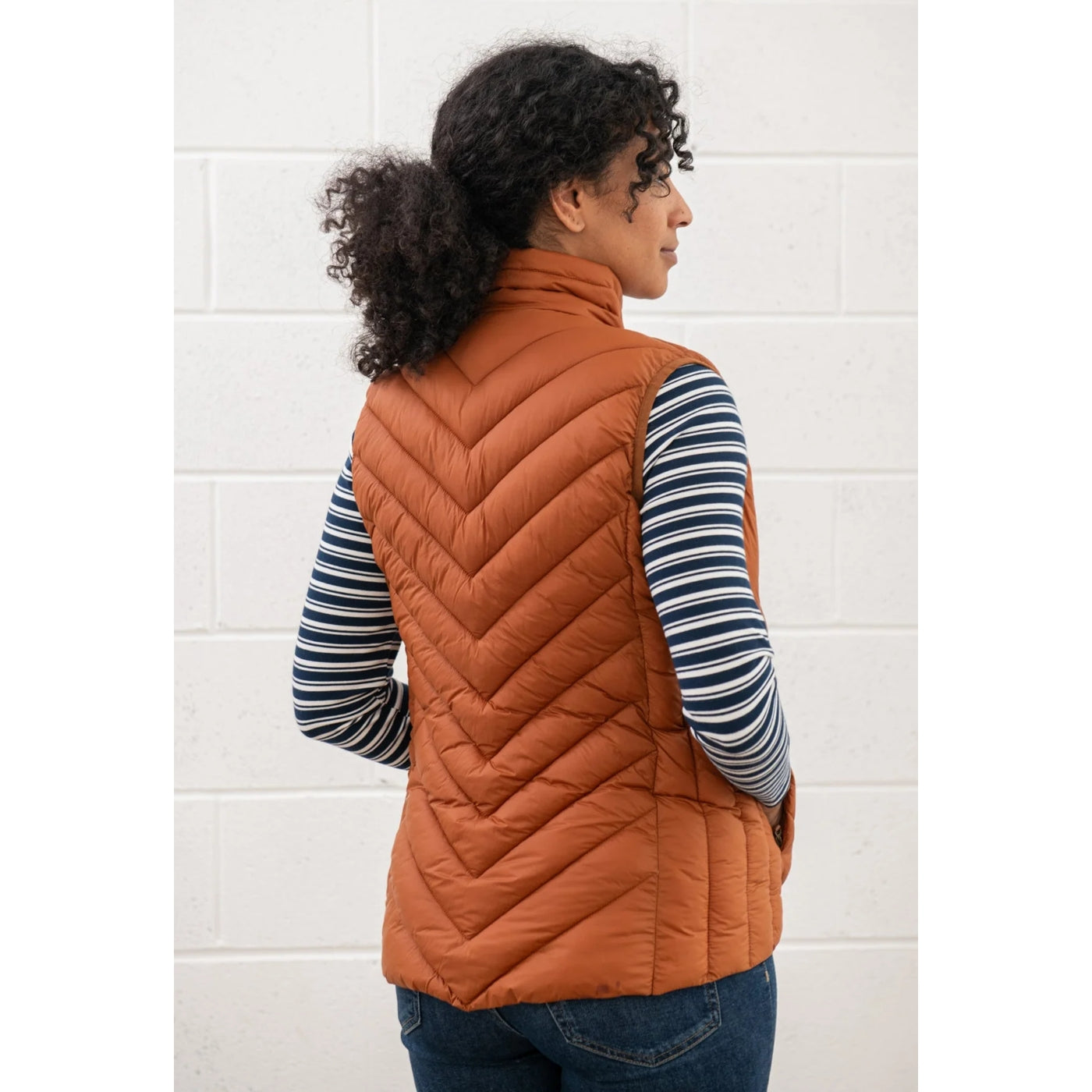 Woman wearing a Rust Orange Lighthouse padded gilet over a striped shirt against a white tiled wall.