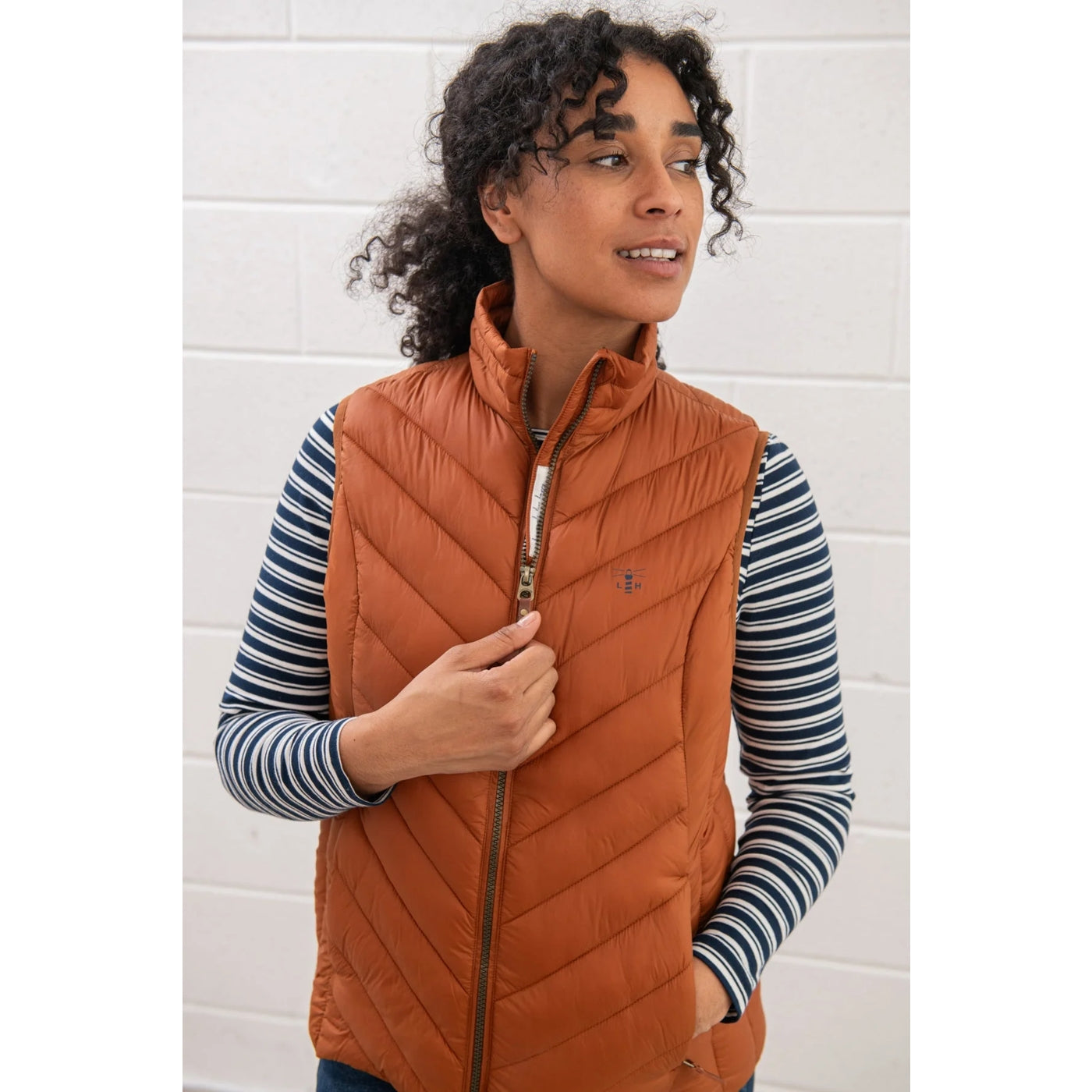 Woman wearing a Lighthouse rust orange puffer gilet over a striped shirt against a white brick wall.