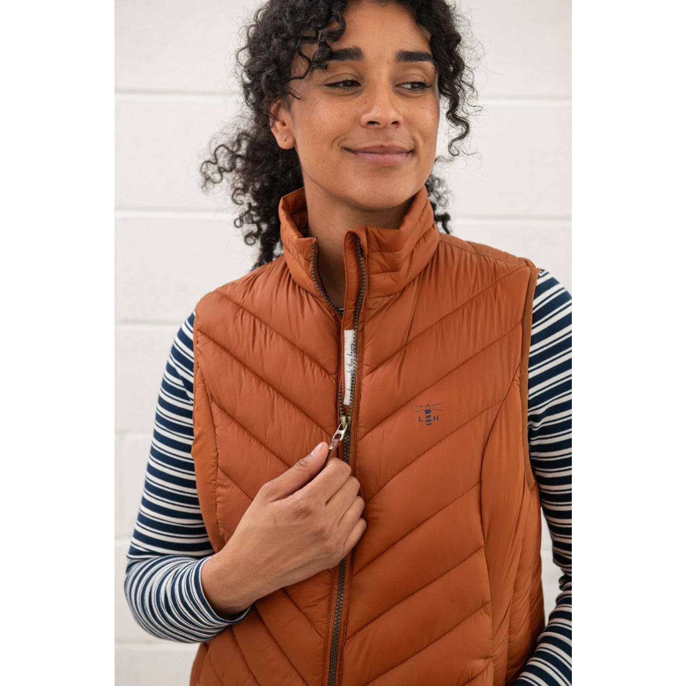 Woman wearing a rust orange Lighthouse Laurel puffer gilet over a striped shirt against a white background
