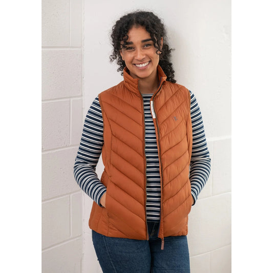 Woman wearing a rust orange Lighthouse Laurel puffer gilet over a striped shirt against a white tiled wall.