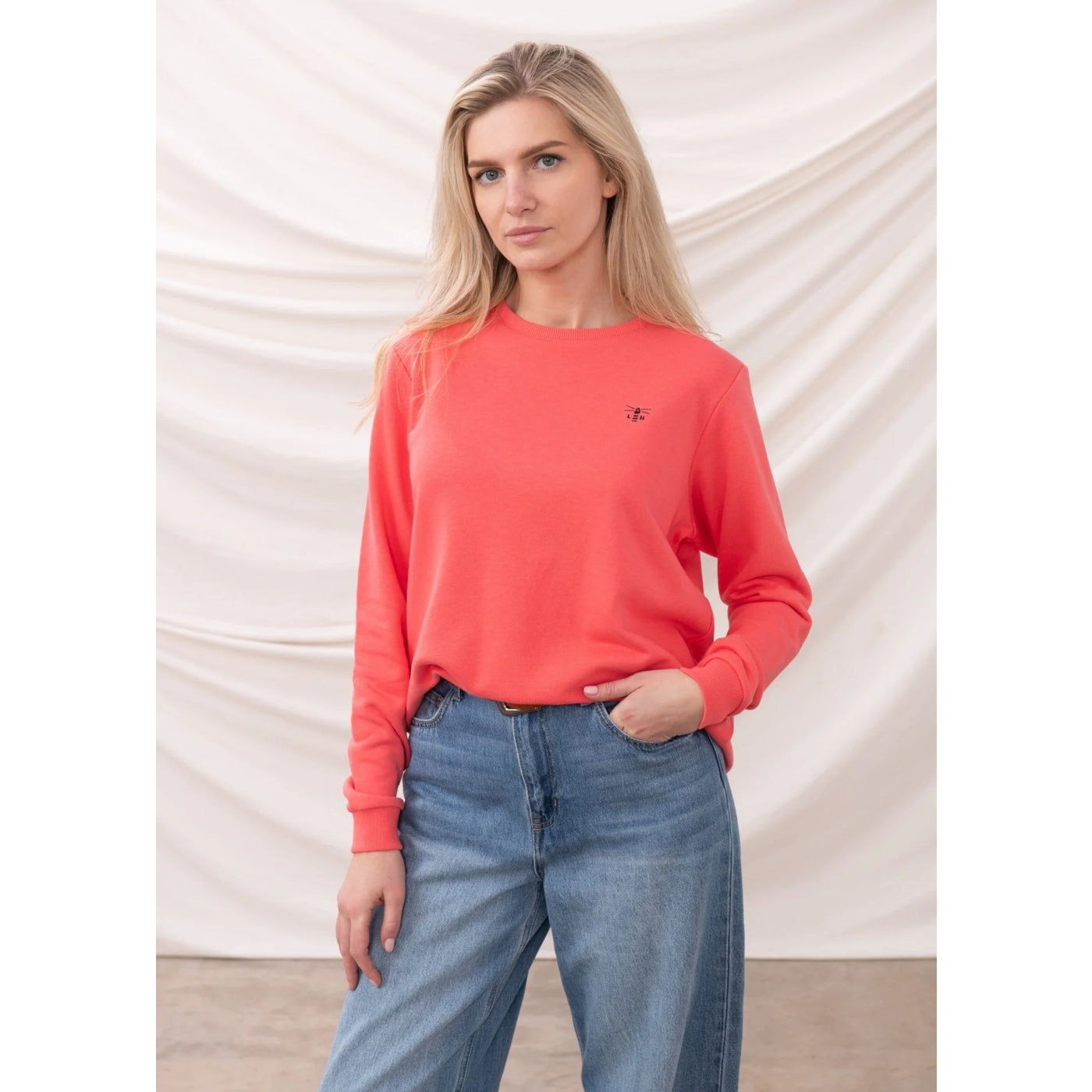 Woman wearing a coral Lighthouse sweatshirt and blue jeans against a plain background