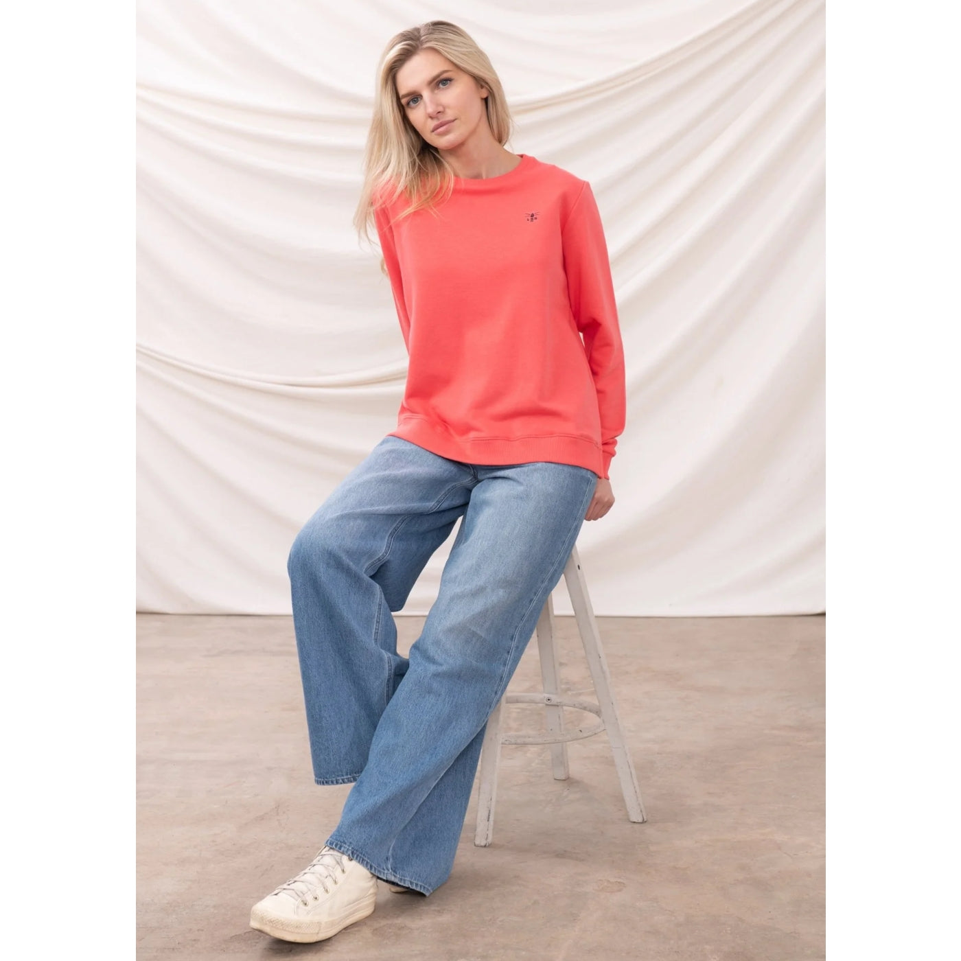 Woman wearing a coral Lighthouse sweatshirt and blue jeans sitting on a stool against a white backdrop
