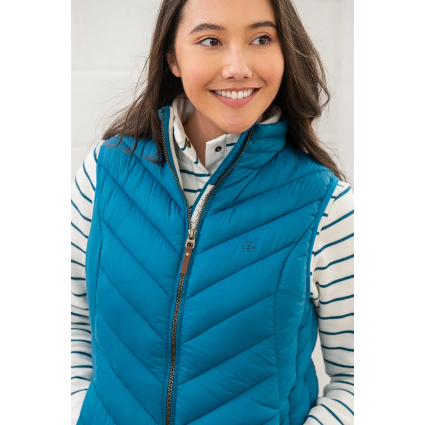 Woman wearing a Lighthouse Laurel teal blue puffer vest over a striped shirt against a white background