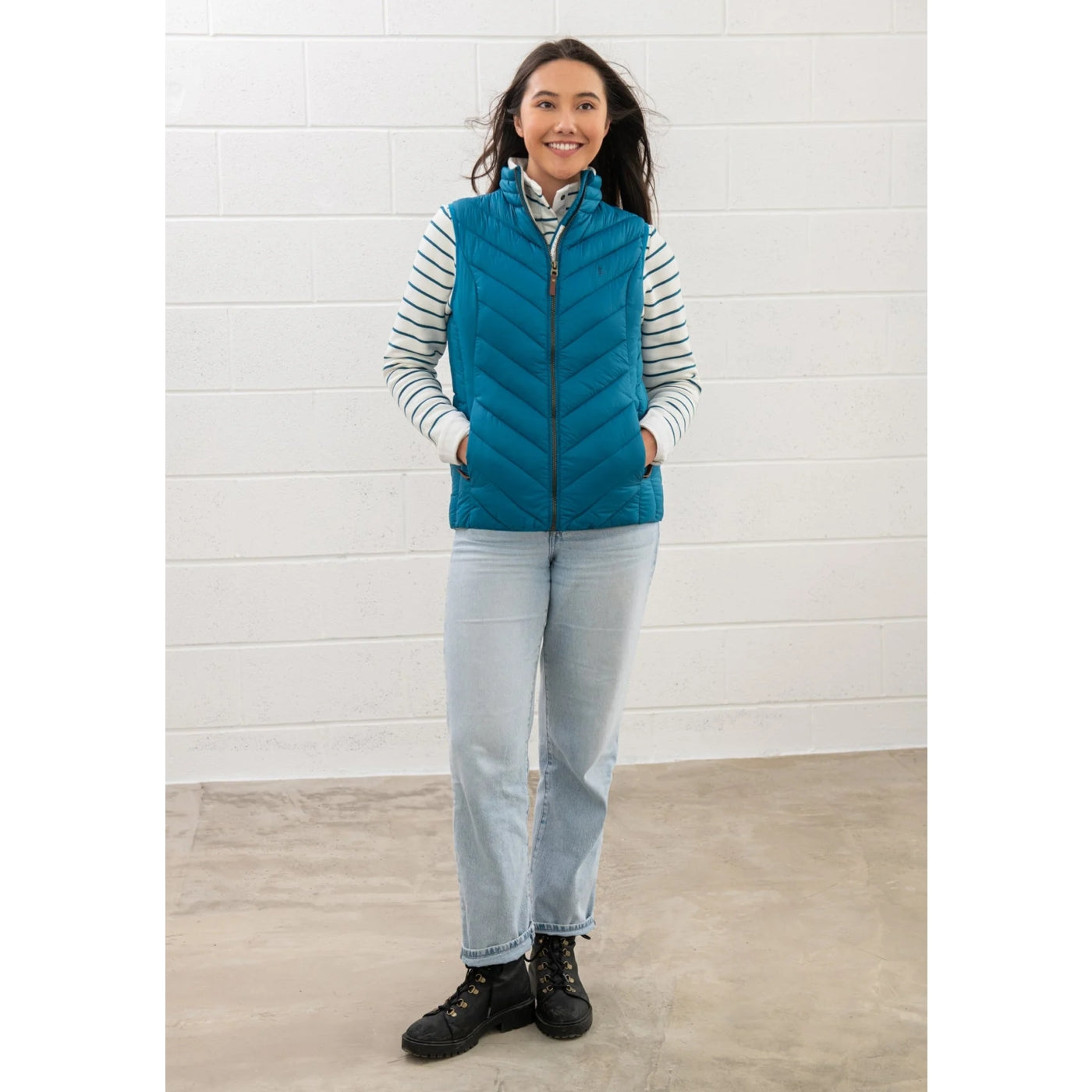 Woman wearing a Lighthouse Laurel puffer gilet in teal blue against a white brick wall.
