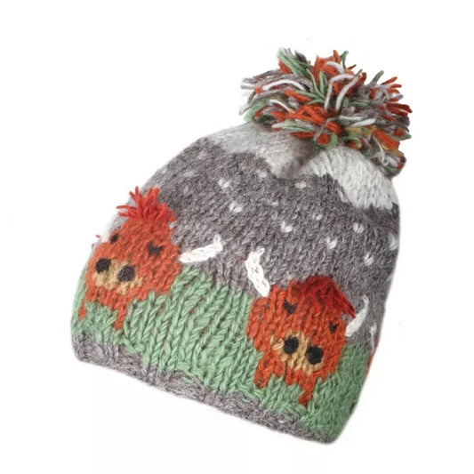 A knitted Pachamama hat in green and grey with a Highland Cow design and pom-pom on a white background