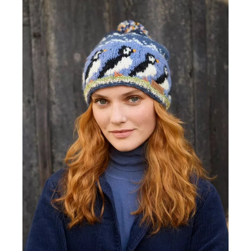 Woman wearing a blue knitted Pachamama beanie hat with puffin design against a wooden background