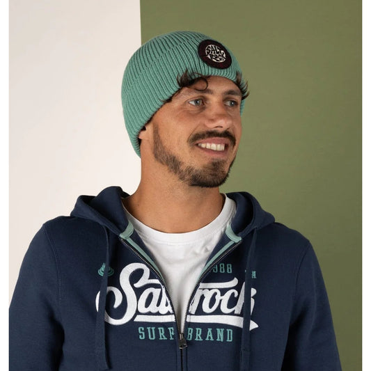 Man wearing a Saltrock SR turquoise knitted beanie and navy hoodie on a green and white background.