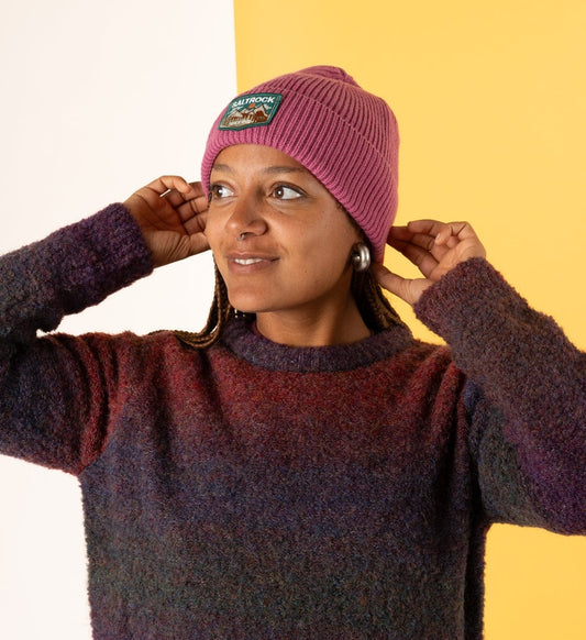 Woman wearing a dark pink Saltrock knitted Uplands beanie with a turn-up hem and mountains logo patch with a sweater against a yellow background