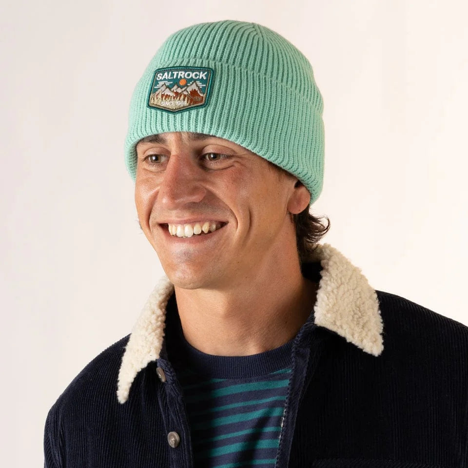 Man wearing a light green Saltrock Uplands beanie with a turn-up hem and mountains logo patch, smiling against a white background