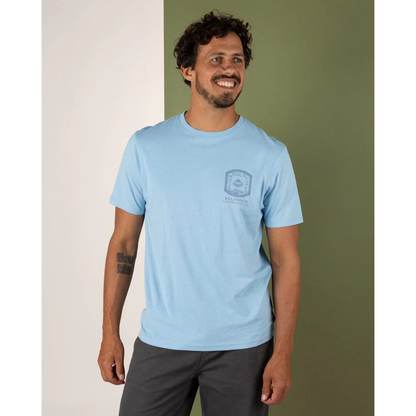Man wearing a light blue t-shirt with a logo against a green wall