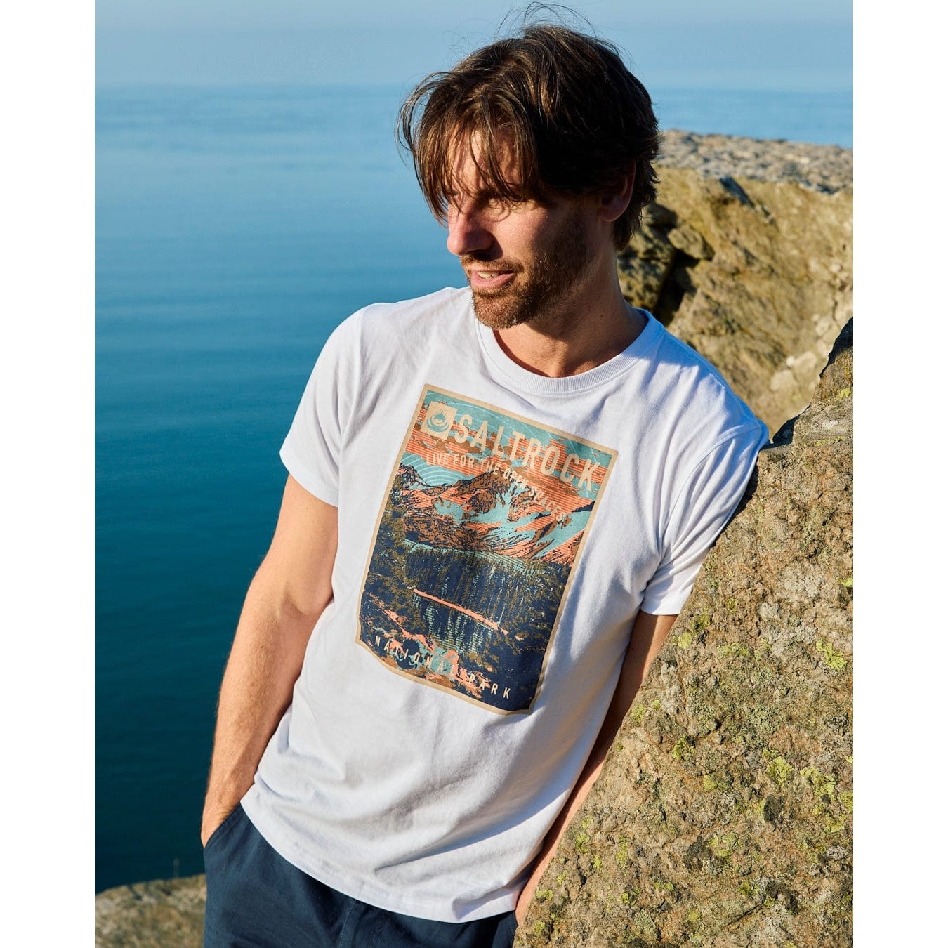 Man wearing a white Saltrock t-shirt with a graphic design standing on a rocky landscape.