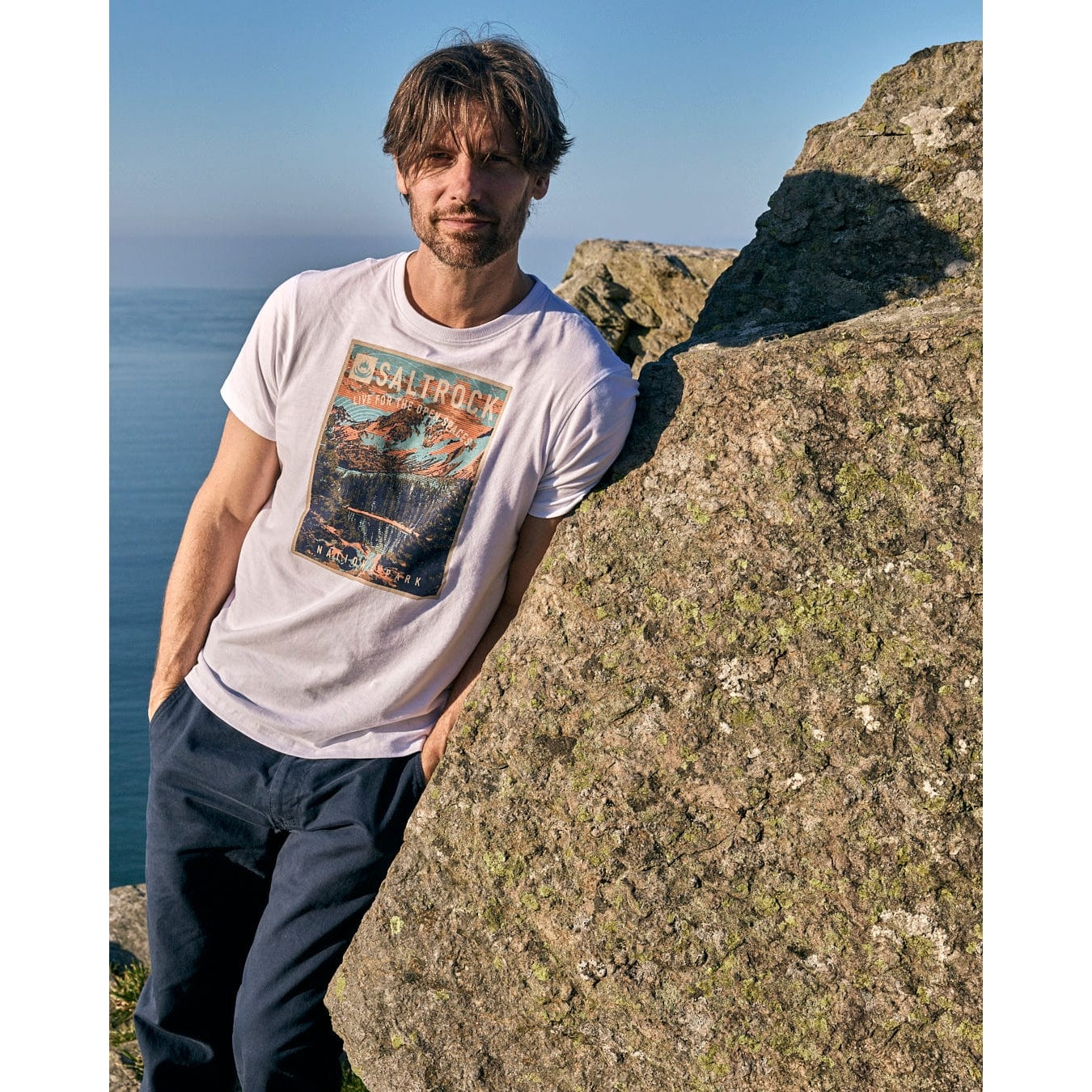 Man wearing a Saltrock t-shirt with a graphic design, standing on a rocky cliff overlooking the ocean.