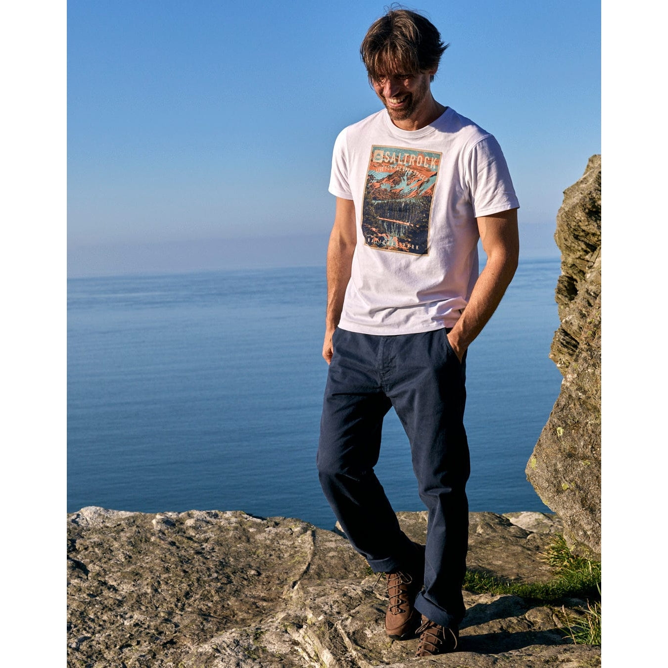 Man wearing a white Saltrock t-shirt with a graphic design, standing on a rocky outcrop by the sea.