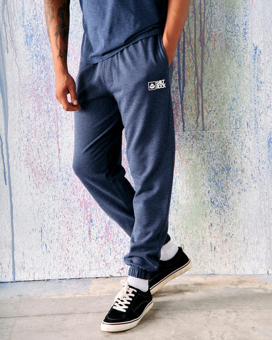Front view of a man wearing dark blue Saltrock Original jogger sweat trousers and sneakers, standing in front of a colourful background.