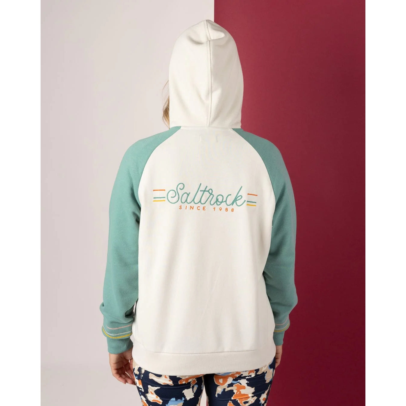 Woman wearing a Saltrock Links Contrast cream and light blue hoodie with 'Saltrock' logo on the back against a red and white background
