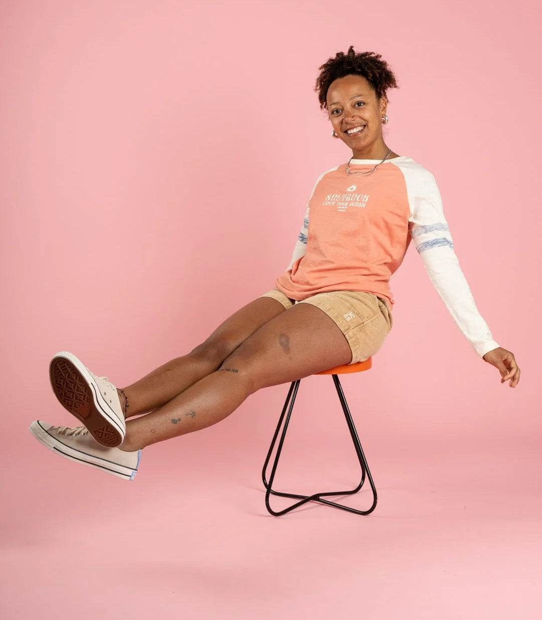 Woman wearing a Saltrock coral and cream t-shirt sitting on a stool against a pink background