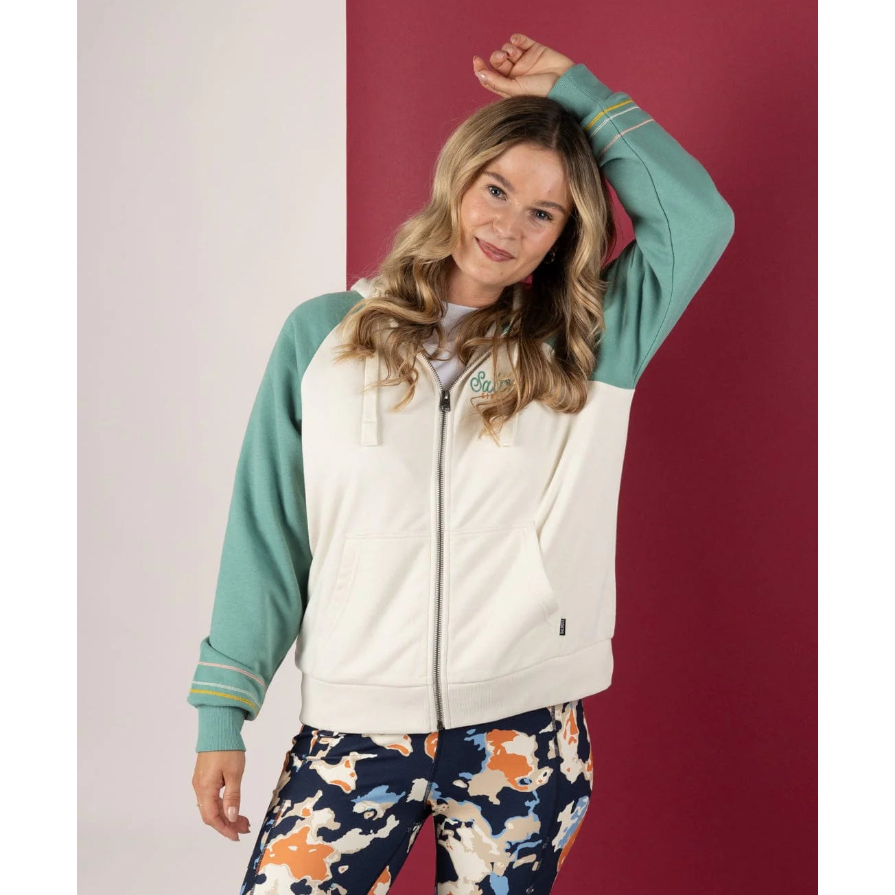 Front view of a woman wearing a Saltrock Links contrast zip hoodie in cream with light blue raglan sleeves on a white and red background.