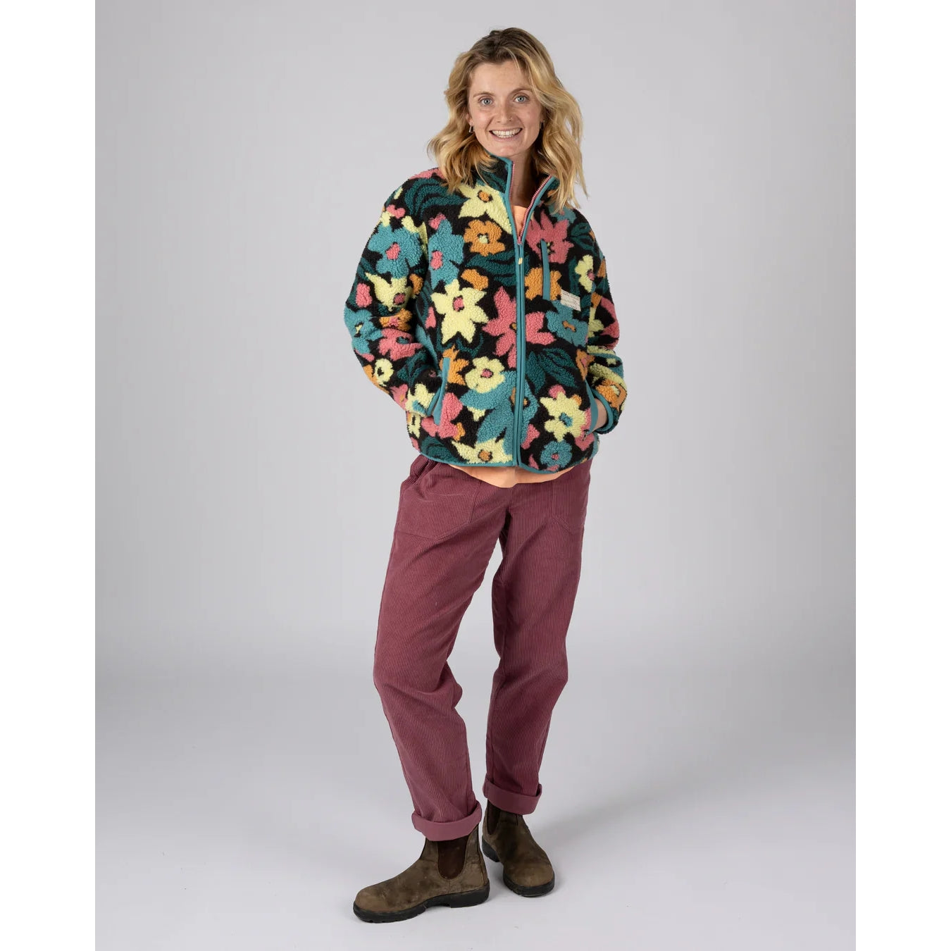 A woman wearing a multicolour floral Saltrock Holidaze zip fleece with dark pink trousers and boots