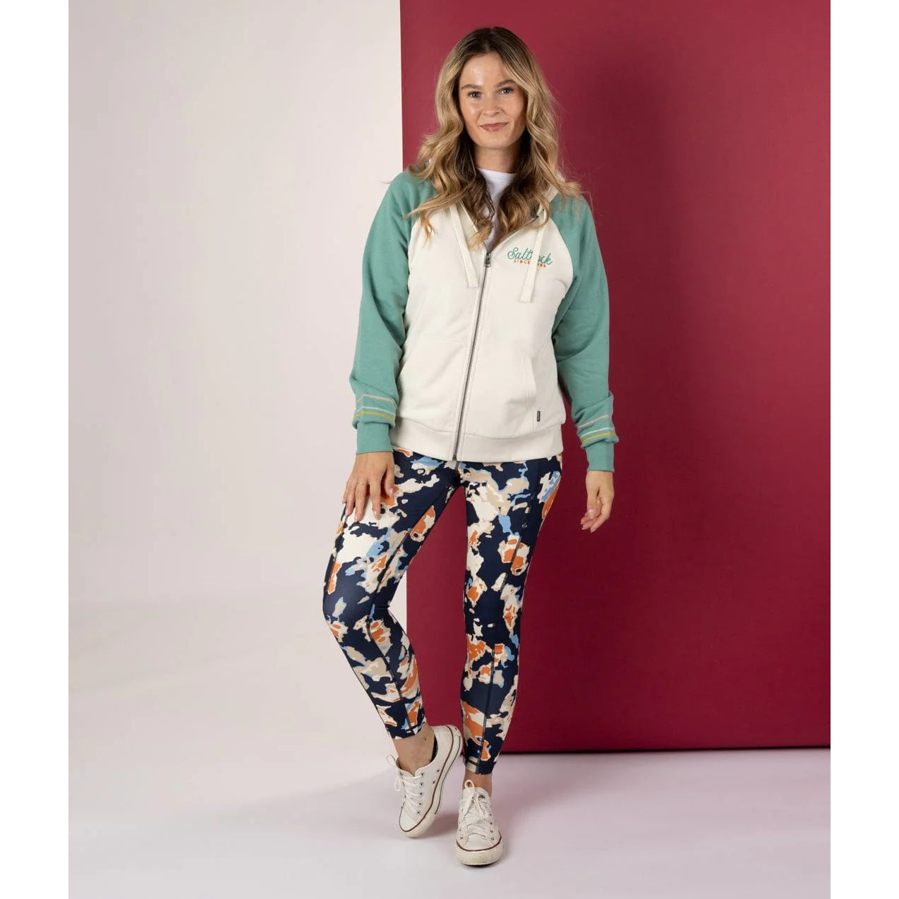 Woman wearing a light blue and cream Saltrock Links Contrast zip hoodie with floral leggings against a red and white background