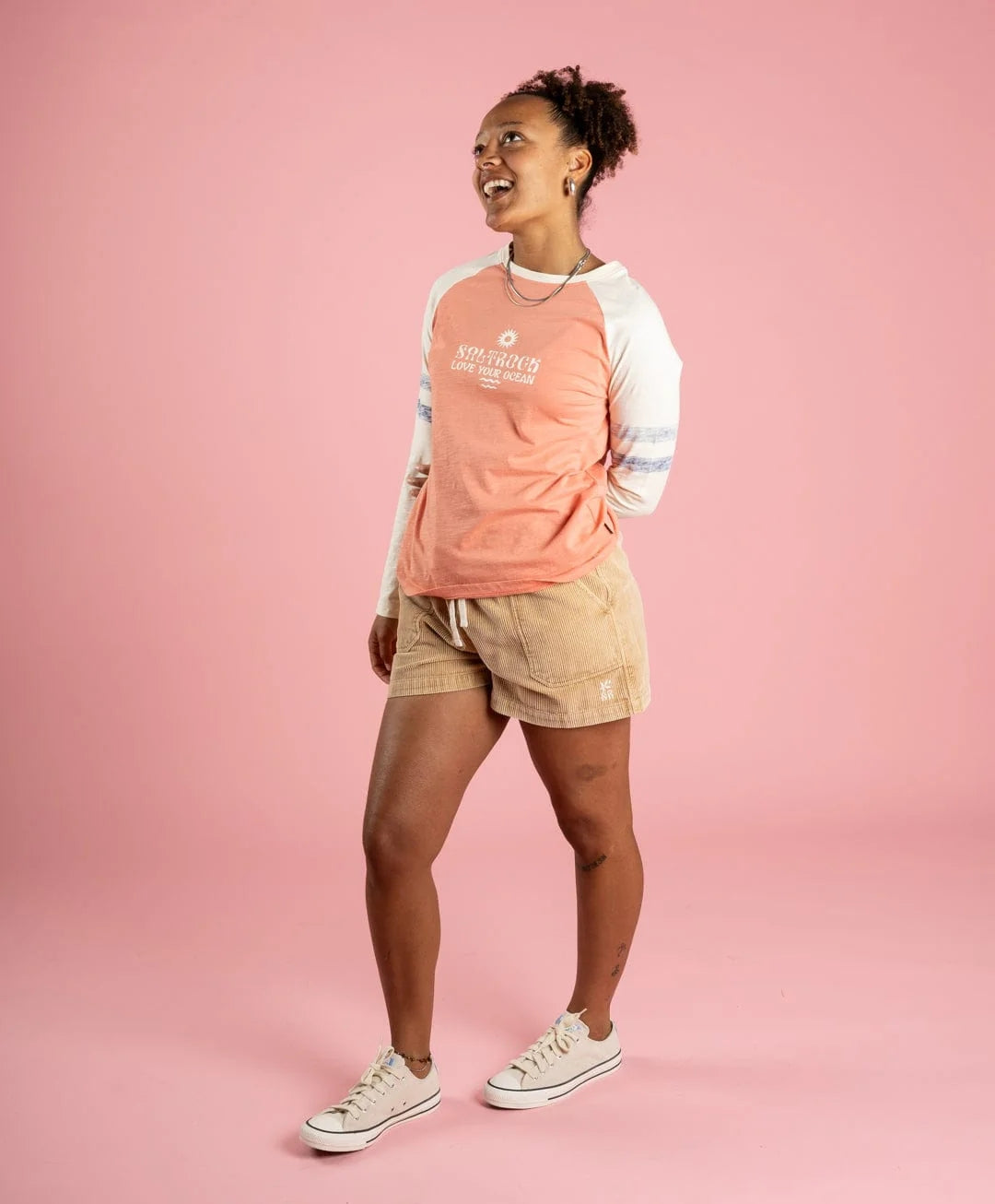 Person wearing a coral and cream long-sleeve t-shirt with beige shorts on a pink background