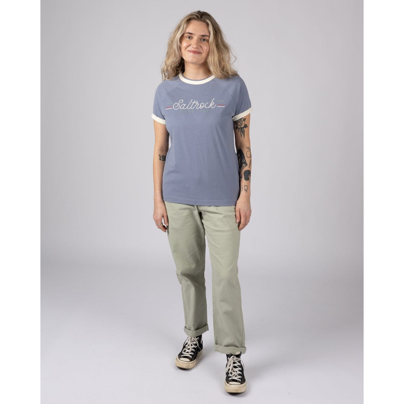 Woman wearing a blue Saltrock Links t-shirt with chest logo and beige trousers on a grey background
