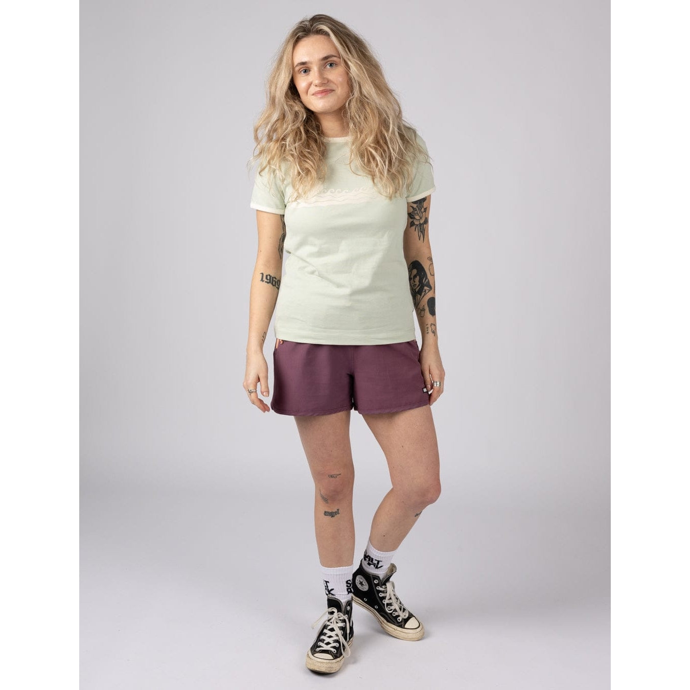 Woman wearing a light green Saltrock Golden Hour short sleeve t-shirt with sunset and sea print across the chest with burgundy shorts and navy sneakers against a plain background
